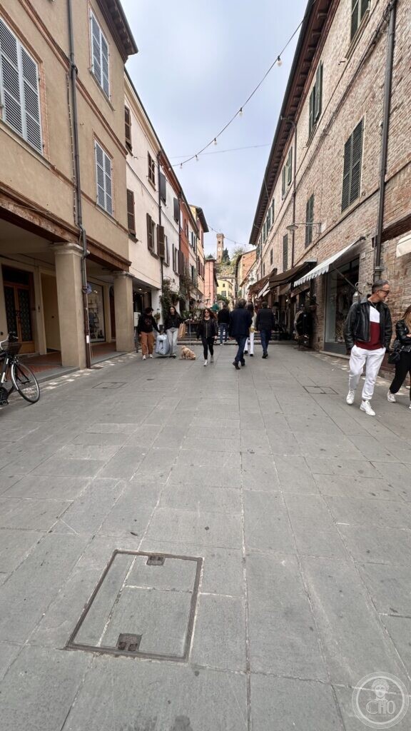 A paved street in the old town of Santarcangelo di Romagna between porticoes and shopfronts, with festive lights strung overhead and the hill town at the far end