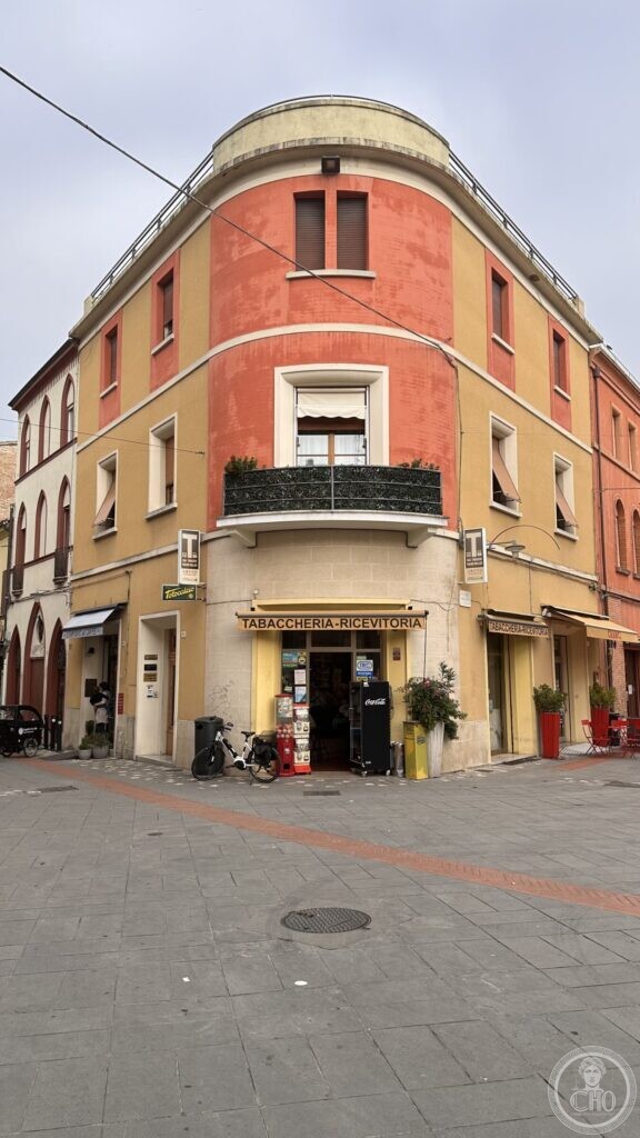 A rounded 1930s corner building in ochre and red in Santarcangelo di Romagna, with two black and white enamel T signs of the state tobacco monopoly and an awning reading TABACCHERIA-RICEVITORIA