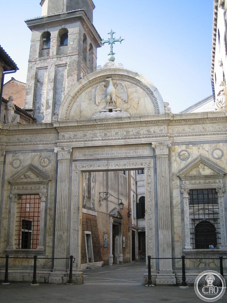 Renaissance marble screen with a central arched portal, a lunette carved with an eagle, pilasters and a cross on top, seen from the campo