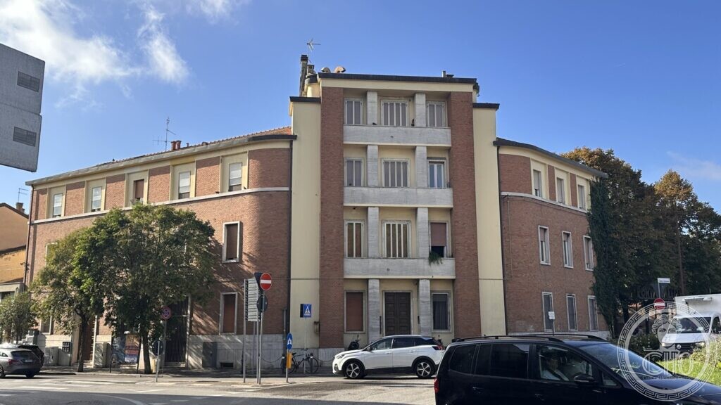 A four-storey block with concrete bands beside a lower brick wing, on a street corner