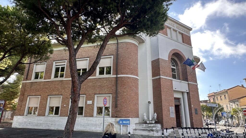 The building seen past a stone pine, brick above a white render plinth, with the entrance arch at the right