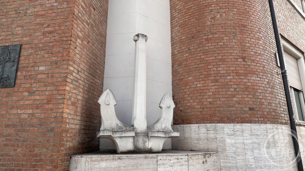 A stylised stone anchor standing against the rounded brick corner of the building, with a bronze relief on the wall beside it