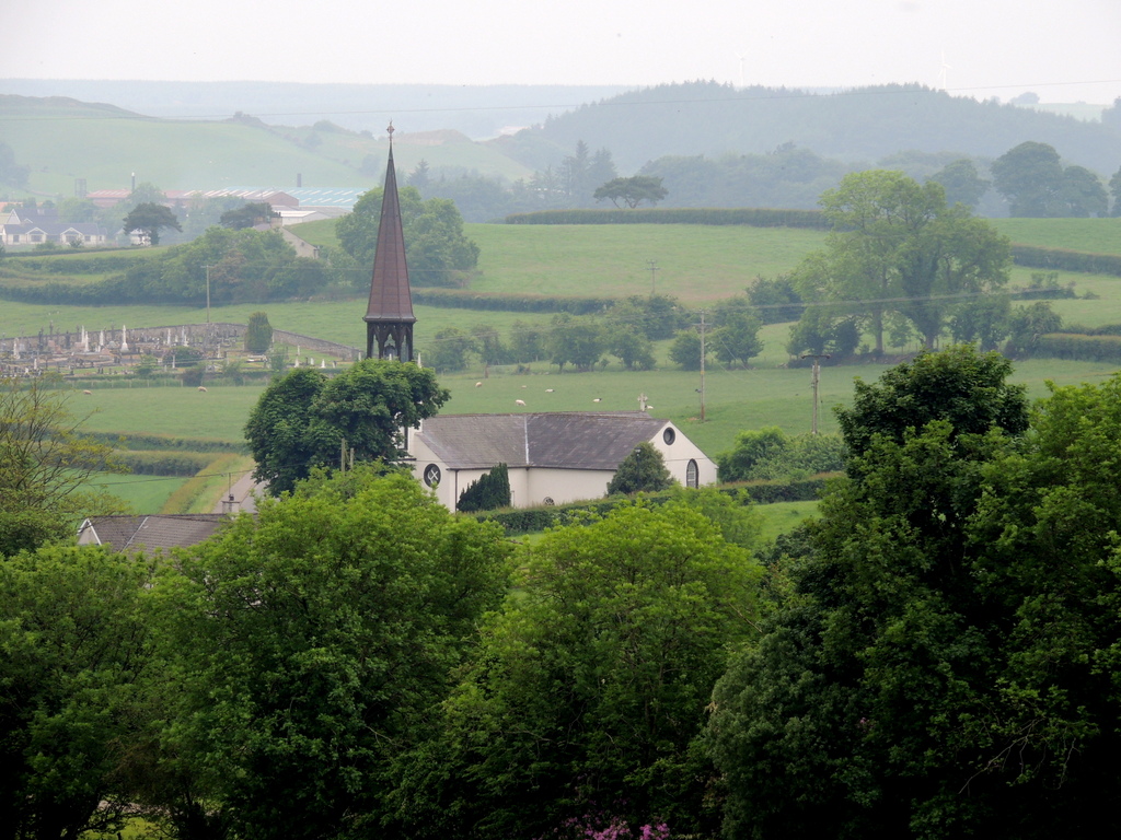 St Eugene's Church at Glenock, a white rendered church with a slate roof and a tall copper-clad spire, among green fields and trees near Newtownstewart