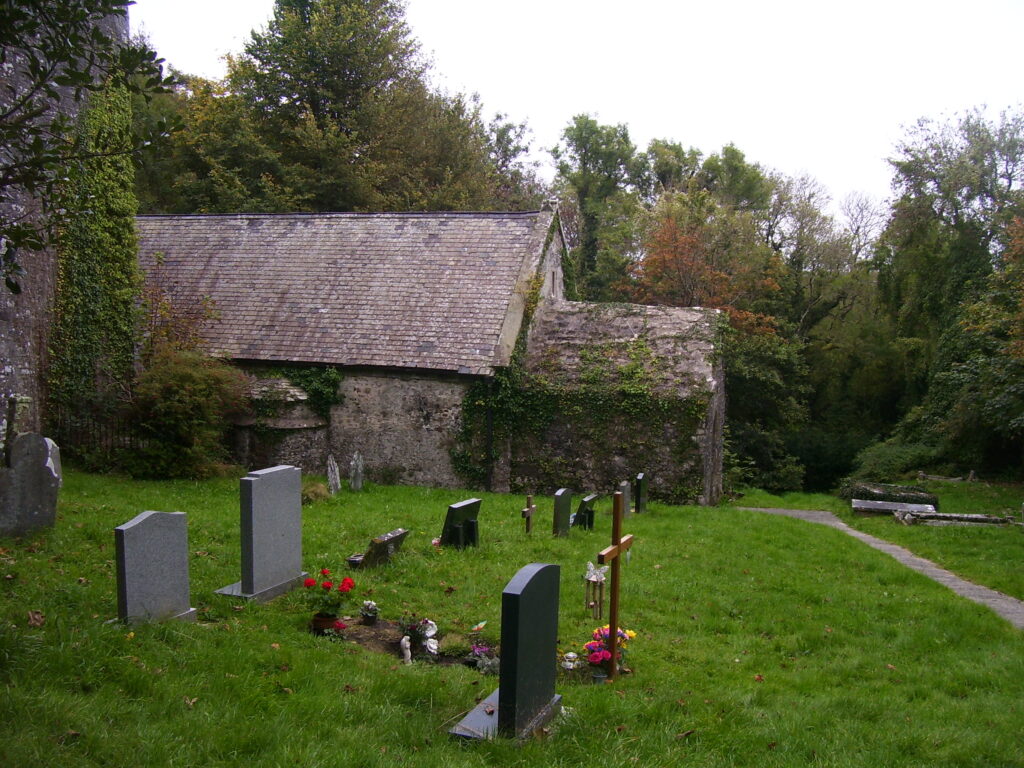 St Lawrence's Church, Gumfreston: a low stone church with a slate roof, its tower covered in ivy at the left, headstones in the grass of the churchyard and a path leading down towards trees