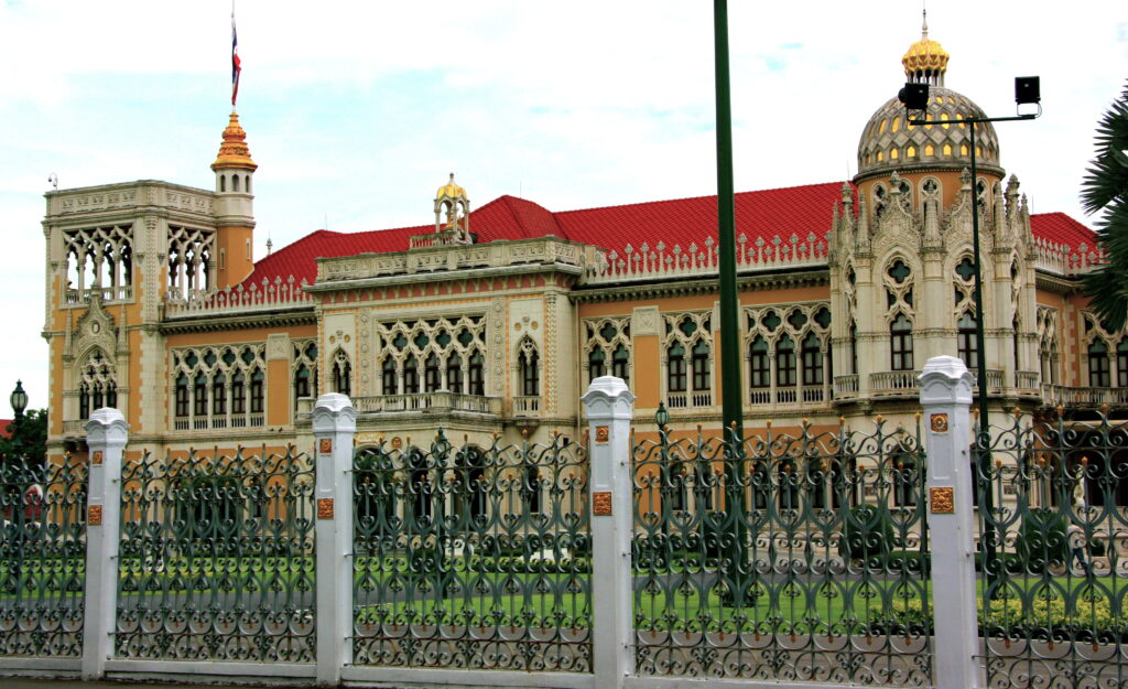 Daylight view of the front of the Thai Khu Fah Building in ochre and cream under a red tiled roof, its upper floor a continuous arcade of pointed Venetian Gothic tracery, behind green railings with gilded panels