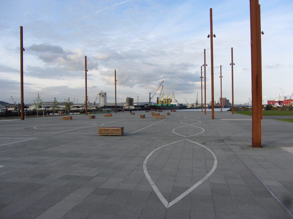 Grey paved slipway in Belfast with white stone outlines, wooden bench blocks and tall rust-brown lamp posts, looking towards the water and the port cranes