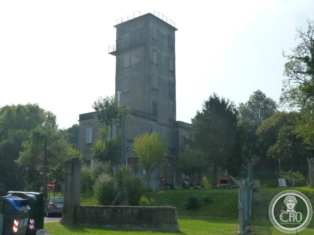 A tall square grey concrete tower with small windows and railings on its top, rising from a lower building among trees and a lawn