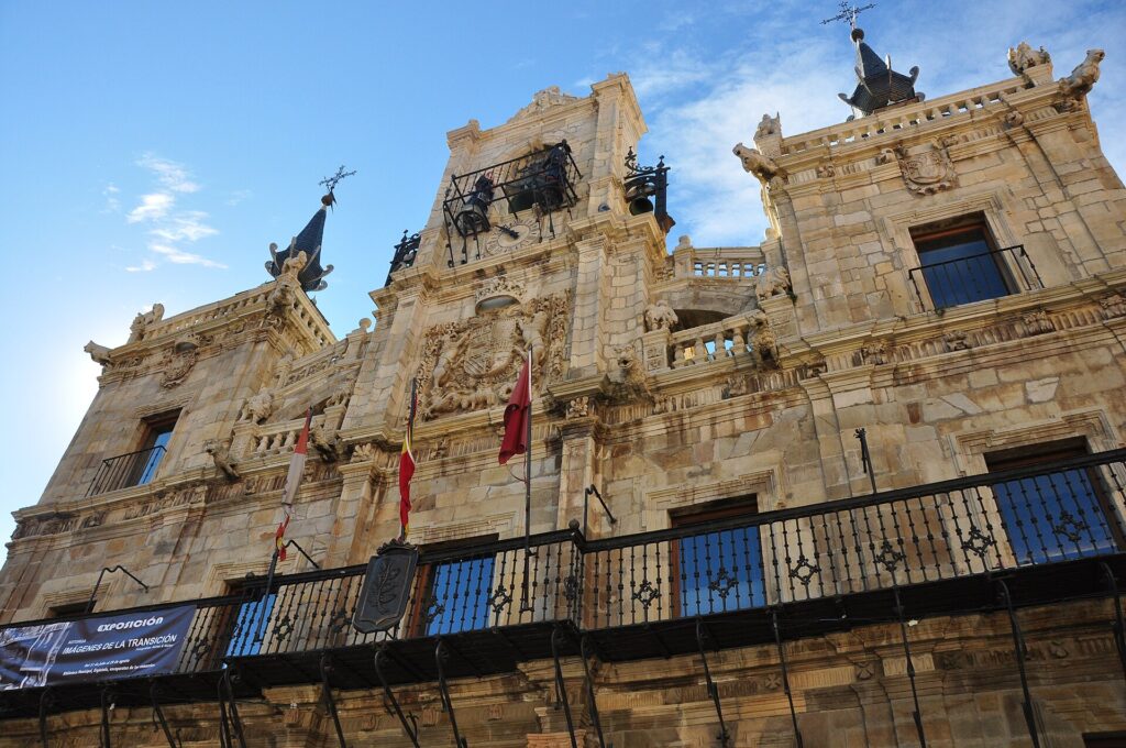 Upward view of the Baroque stone front of Astorga town hall, with a long iron balcony, carved coats of arms, a central bell frame and two towers crowned by slate spires