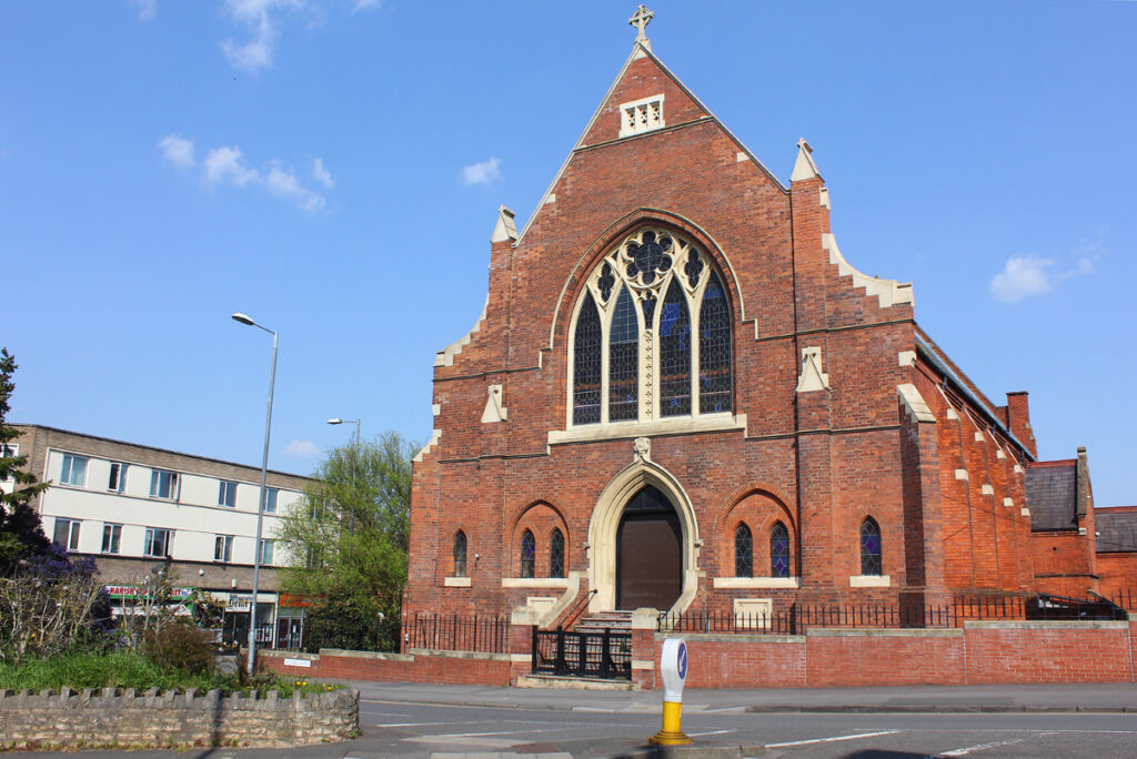 Red-brick gabled front of Trinity Hall, Swindon, with a large traceried pointed window, a stone-framed doorway at the top of steps and pinnacled buttresses