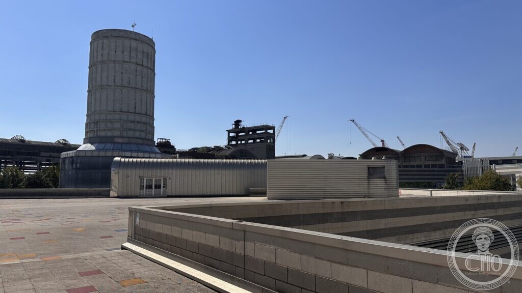 A cooling tower and the cranes of the Marghera industrial port rising behind the low buildings of the park