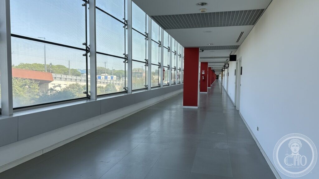A long corridor with a fully glazed wall on one side and red doors on the other
