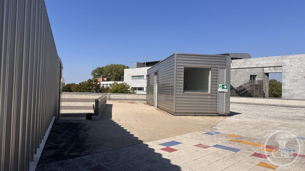 A small metal-clad prefabricated pavilion standing on a paved terrace between larger buildings