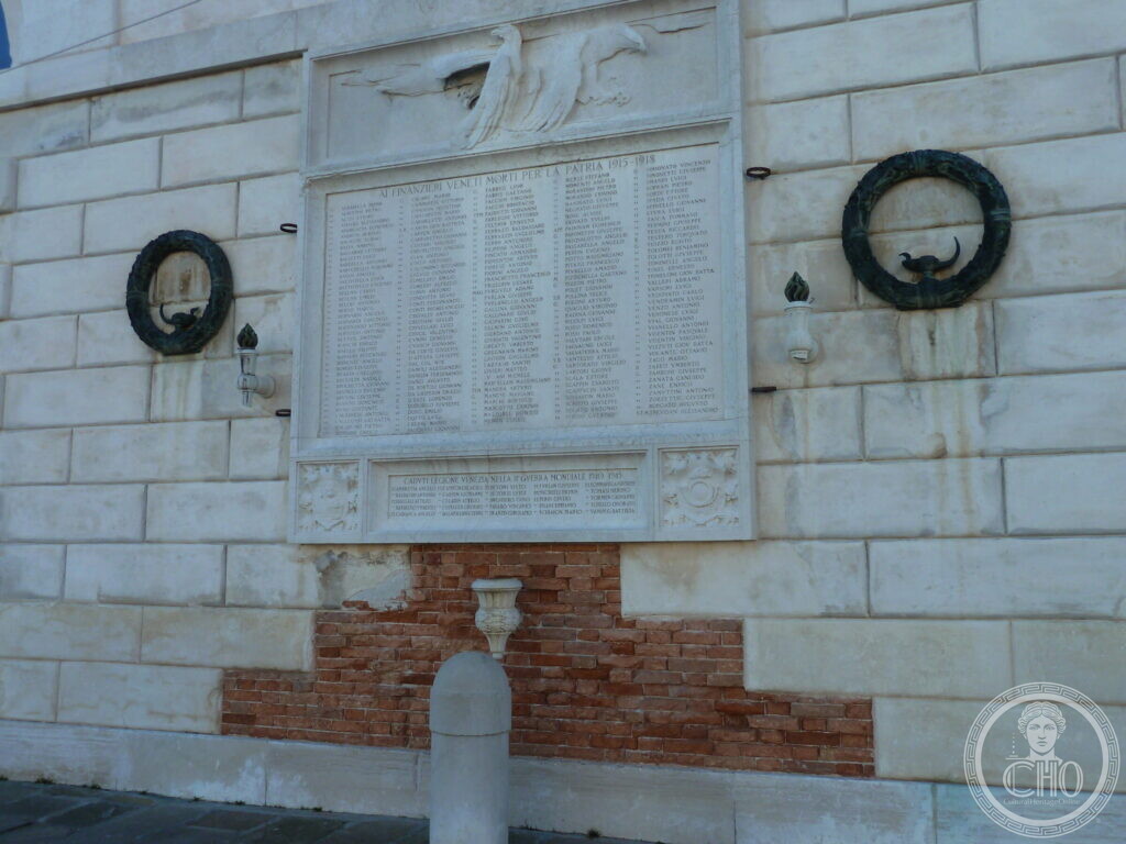 The memorial tablet seen against the whole white stone wall, with exposed brick at its base