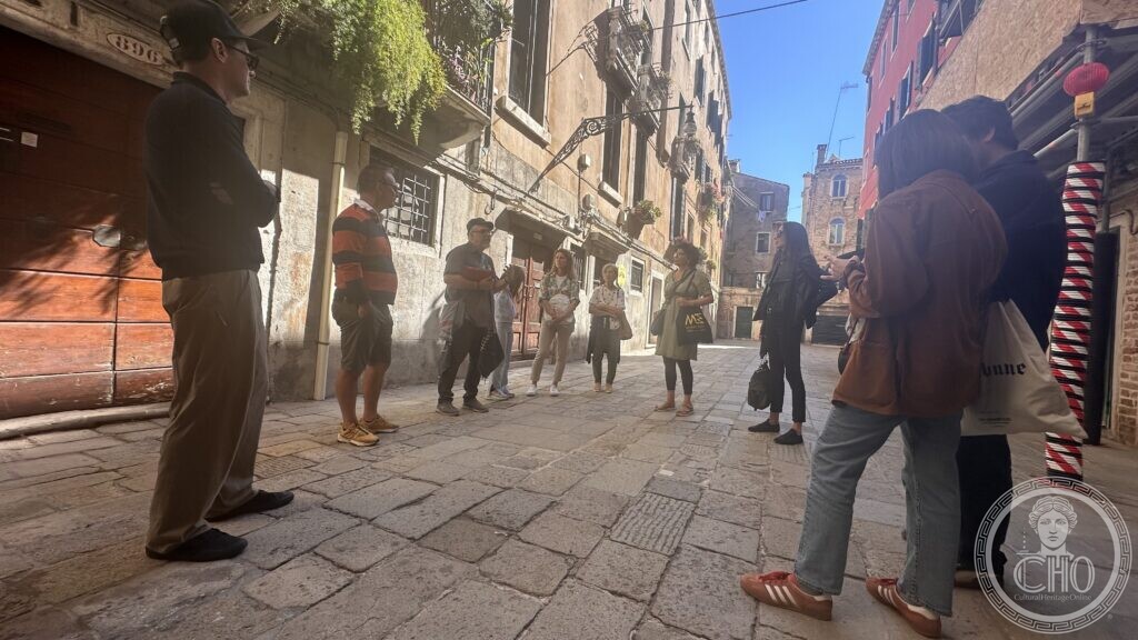 A group of walkers in a Venetian calle with old houses, listening to their guide