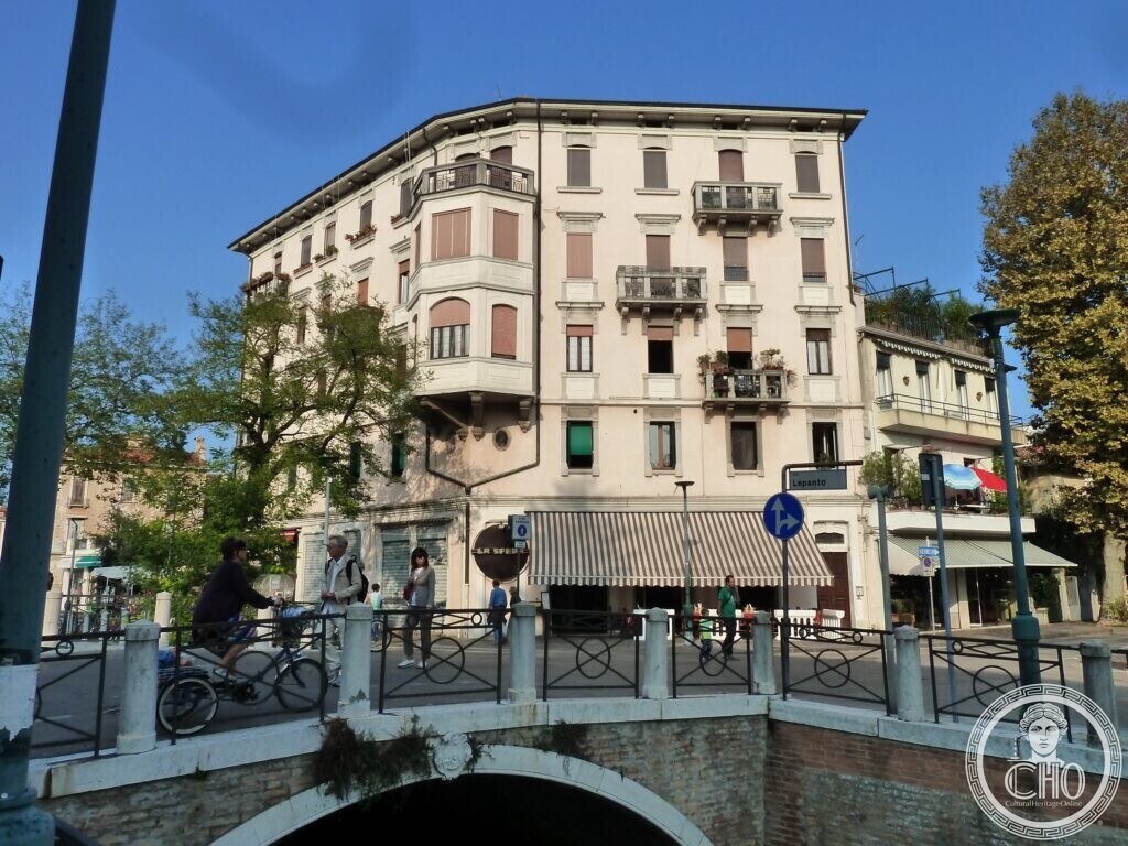 A tall pale corner apartment block with bay windows, balconies and a striped shop awning at street level, seen across a small stone bridge over a canal
