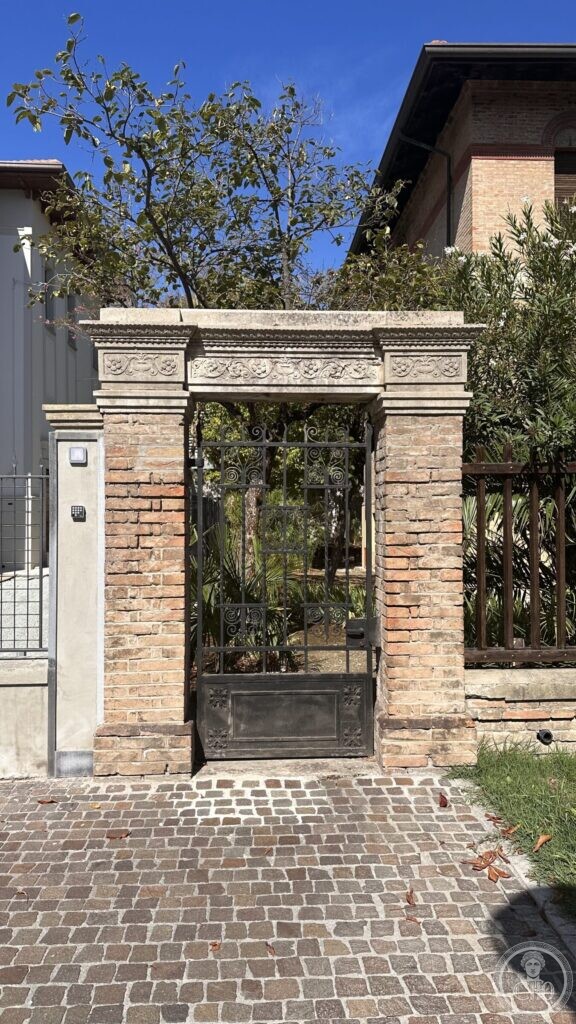 A brick and stone pedestrian gateway with a carved lintel of festoons, closed by an iron gate