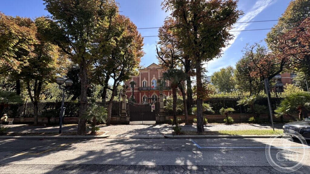 The villa at the end of a line of trees, seen from the opposite pavement of the avenue