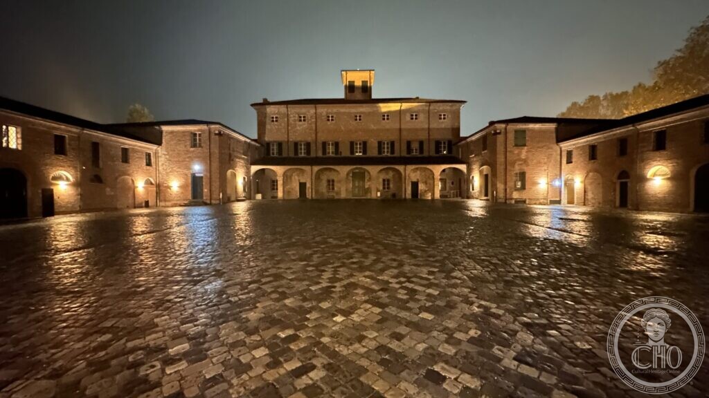 A large cobbled courtyard surrounded by brick ranges with arcades, wet and lit at night