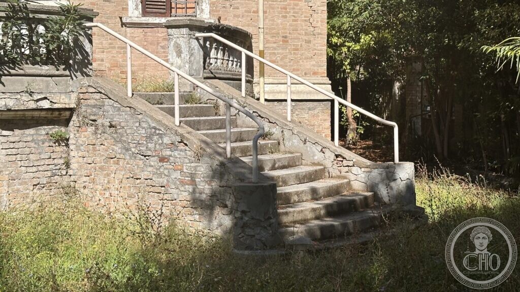 Stone steps with a balustrade and a modern steel handrail, the render fallen away and weeds in the joints