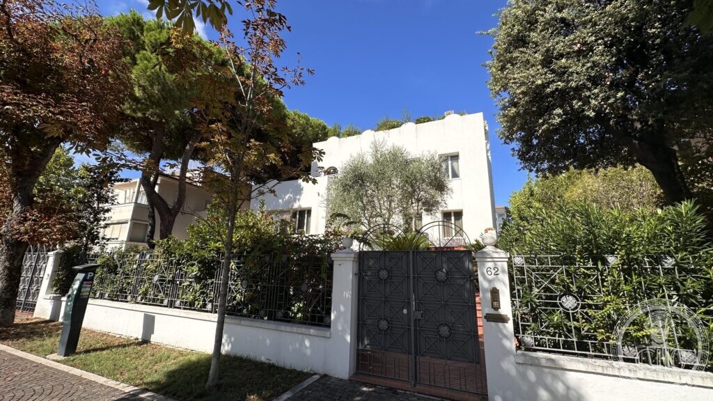 A white modernist villa behind a wrought-iron gate marked 62, with pines and oleanders along the pavement