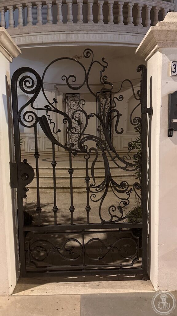 A wrought-iron pedestrian gate filled with Art Nouveau scrolls and tendrils, between rendered piers