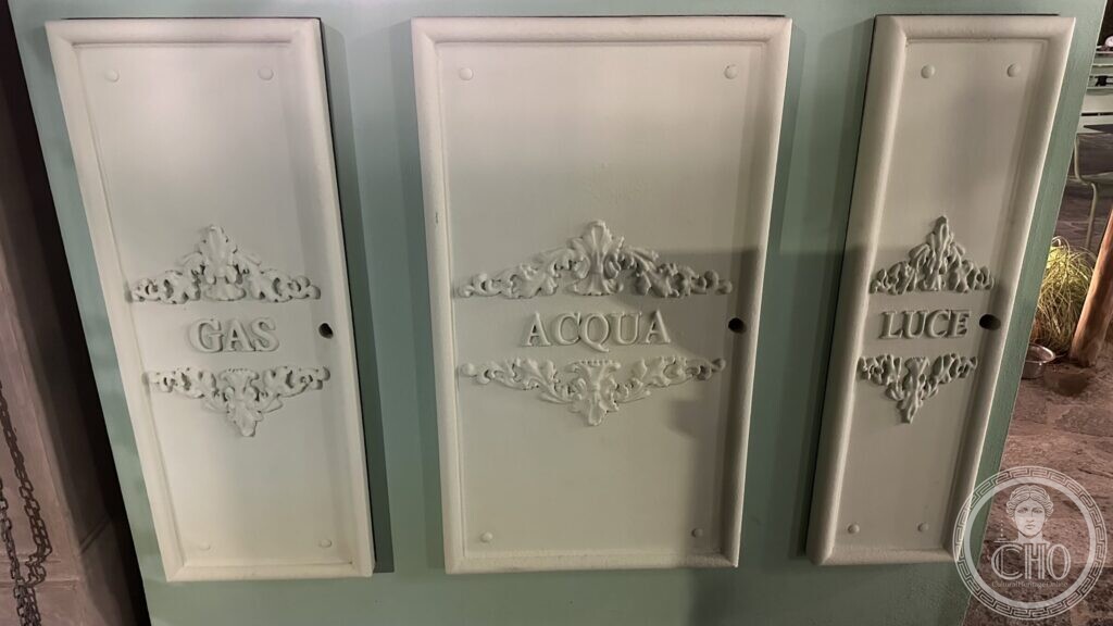 Three cast-iron doors in a green wall, marked GAS, ACQUA and LUCE, each with moulded foliage