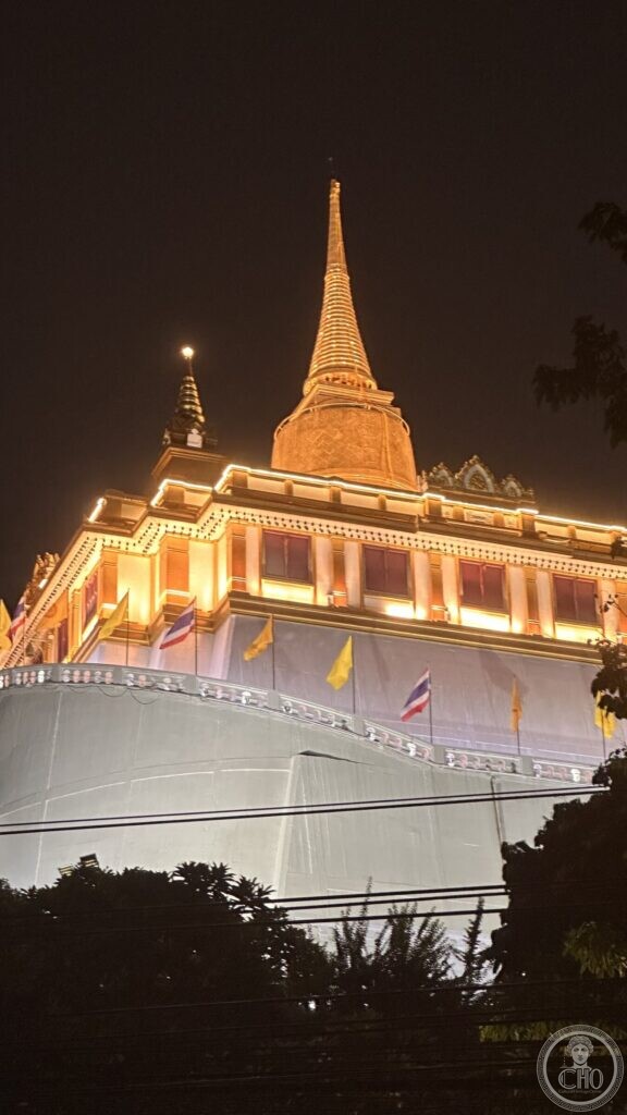 The gilded chedi of Wat Saket lit at night above the white drum of the Golden Mount, with scaffolding sheeting and flags on the terrace