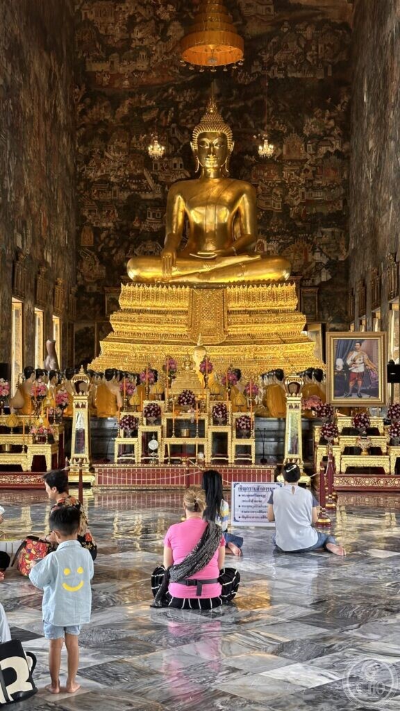 Families sitting on the marble floor of the hall at Wat Suthat facing the gilded Buddha, with a printed notice between them and the altars