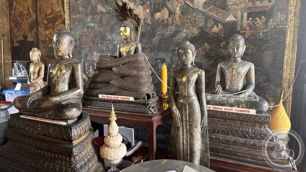 Small dark bronze Buddha images patched with gold leaf, standing and seated on wooden tables at Wat Suthat, with a printed Thai notice asking visitors not to apply gold leaf, pour water or place flowers
