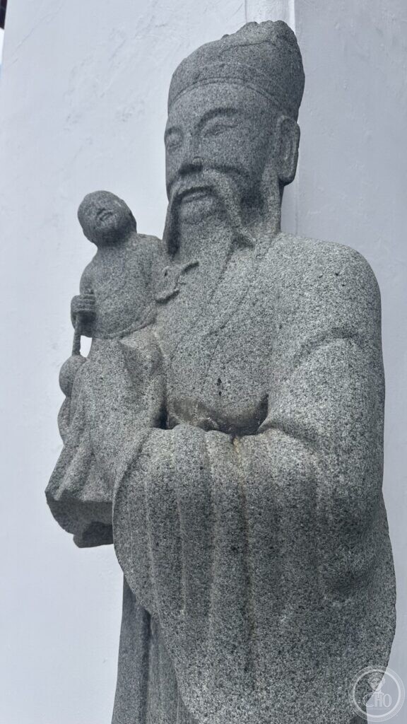 A grey granite Chinese figure of a bearded official in long robes and a tall soft cap, holding a small child on one arm, against a white wall at Wat Suthat