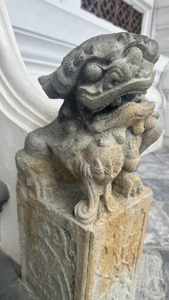 A weathered Chinese granite guardian lion at Wat Suthat, a forepaw on a carved ball, on a pedestal with shallow relief