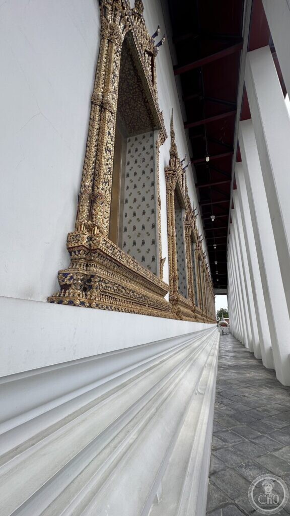 Gilded and inlaid window frames along the white outer wall of the ordination hall at Wat Suthat, seen down the colonnade