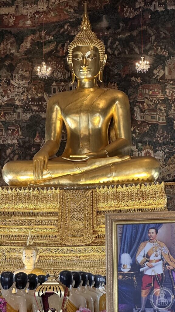 Close view of the gilded bronze Phra Buddha Trilokachet, with cut-glass chandeliers, the painted wall behind, a framed royal portrait and a row of disciple figures below