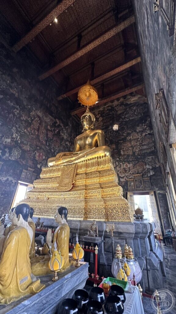 The gilded bronze Phra Buddha Trilokachet on its tall tiered base in the ordination hall of Wat Suthat, under a ribbed golden parasol, with yellow-robed disciple figures in the foreground, hands pressed together