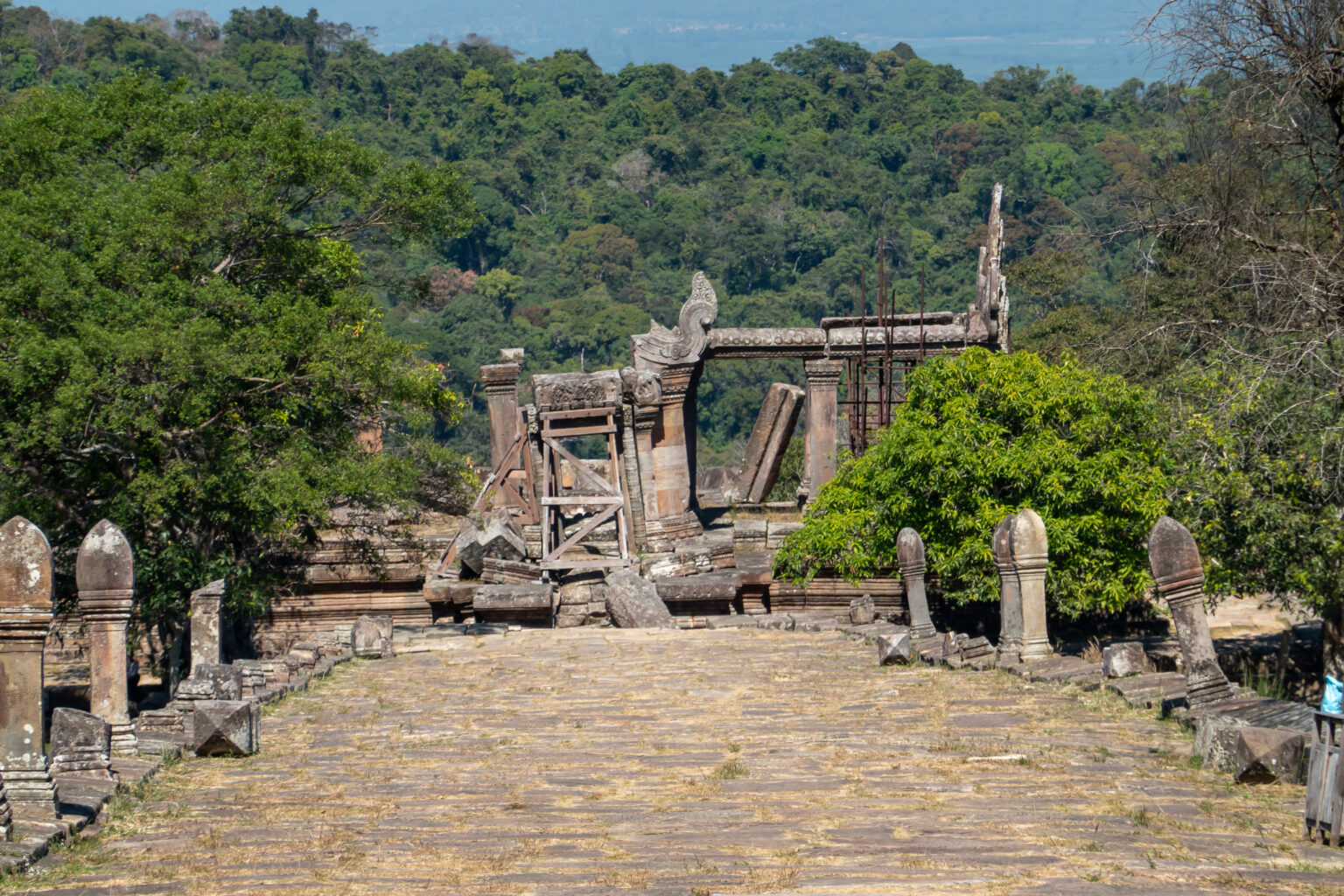 A straight paved processional causeway lined with low boundary posts leading uphill towards a ruined gateway at Preah Vihear