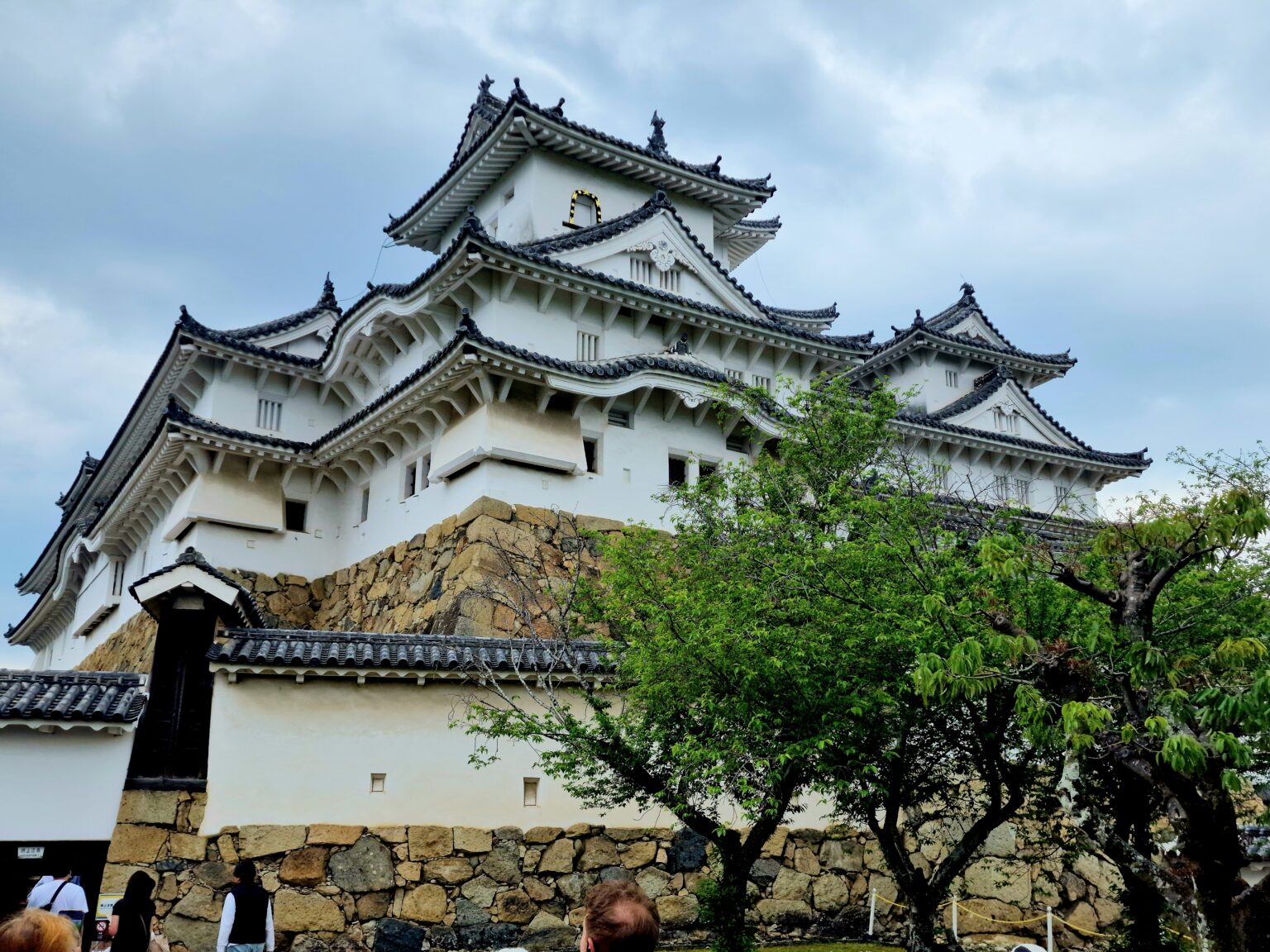 The white keep of Himeji Castle rising in tiers of tiled roofs above a tall sloping stone base, trees in the foreground