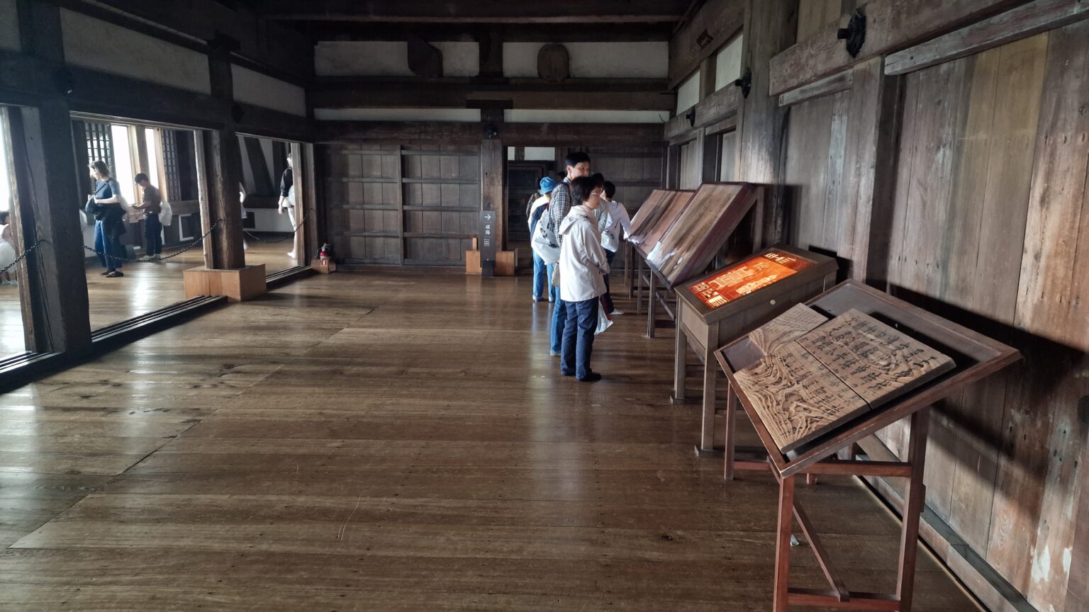 A long bare timber hall inside Himeji Castle with plank floors and whitewashed walls, visitors reading display boards along one side