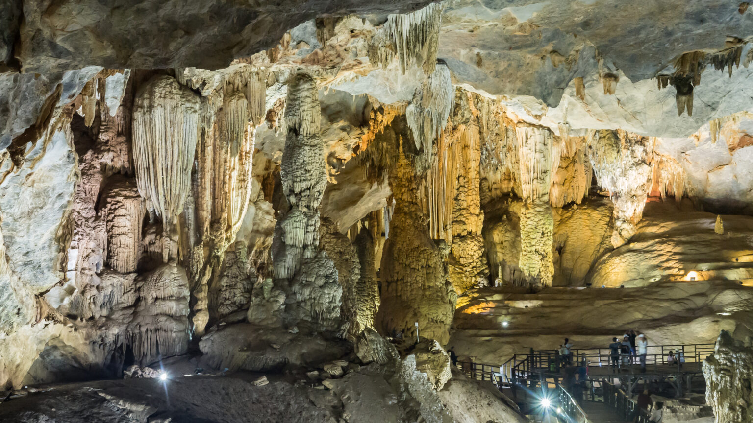 A lit walkway between tall stalagmites and flowstone inside a vast cave chamber