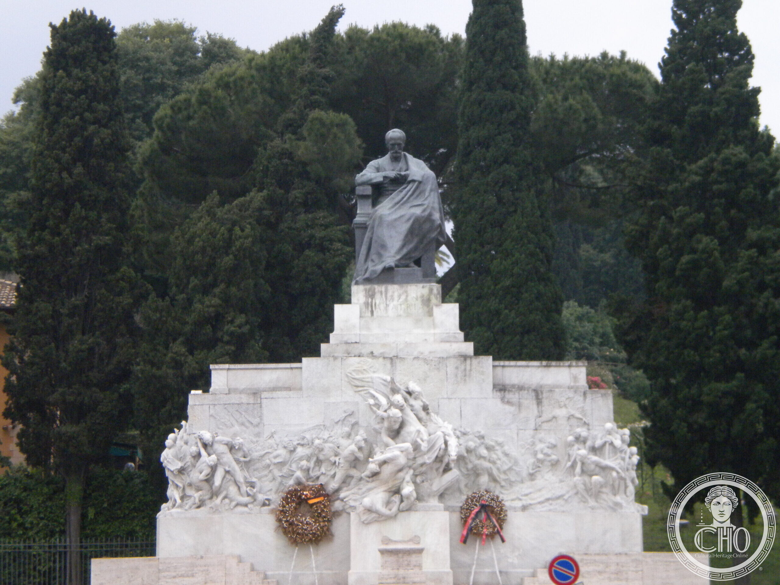 A dark bronze statue of a seated man in a cloak on a tall white marble base carved with figures in relief, two wreaths with ribbons at the foot, cypresses and pines behind