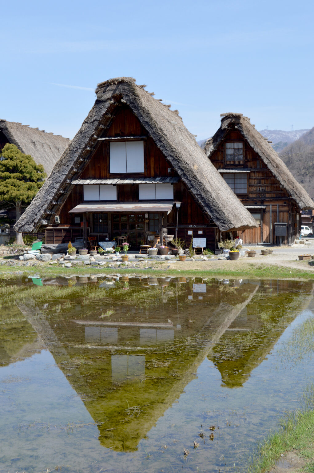 A gasshō farmhouse with a very steep thatched roof and a tall timber gable, reflected in still water in front of it