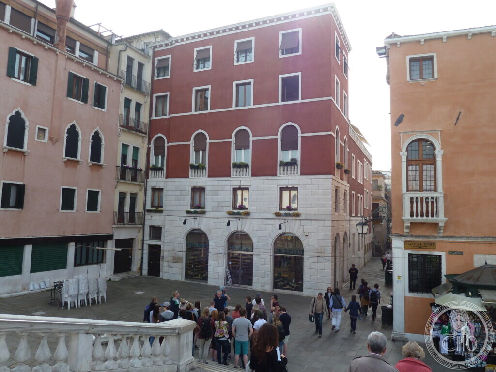 A small Venetian square with a red palazzo behind a stone arcade, crowds of people, and other buildings on either side.