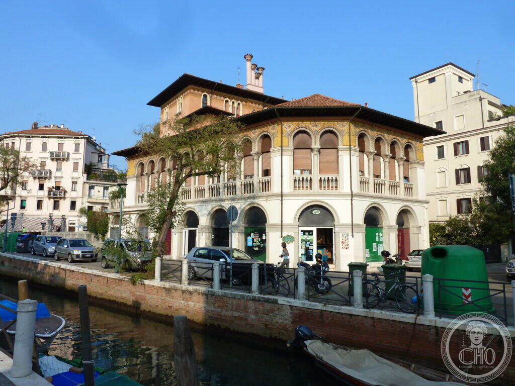 A two-storey corner building in cream plaster by a canal, with round-arched windows on the ground floor, a loggia of arches with slender columns on the first floor and a tower-like block with a tall chimney behind