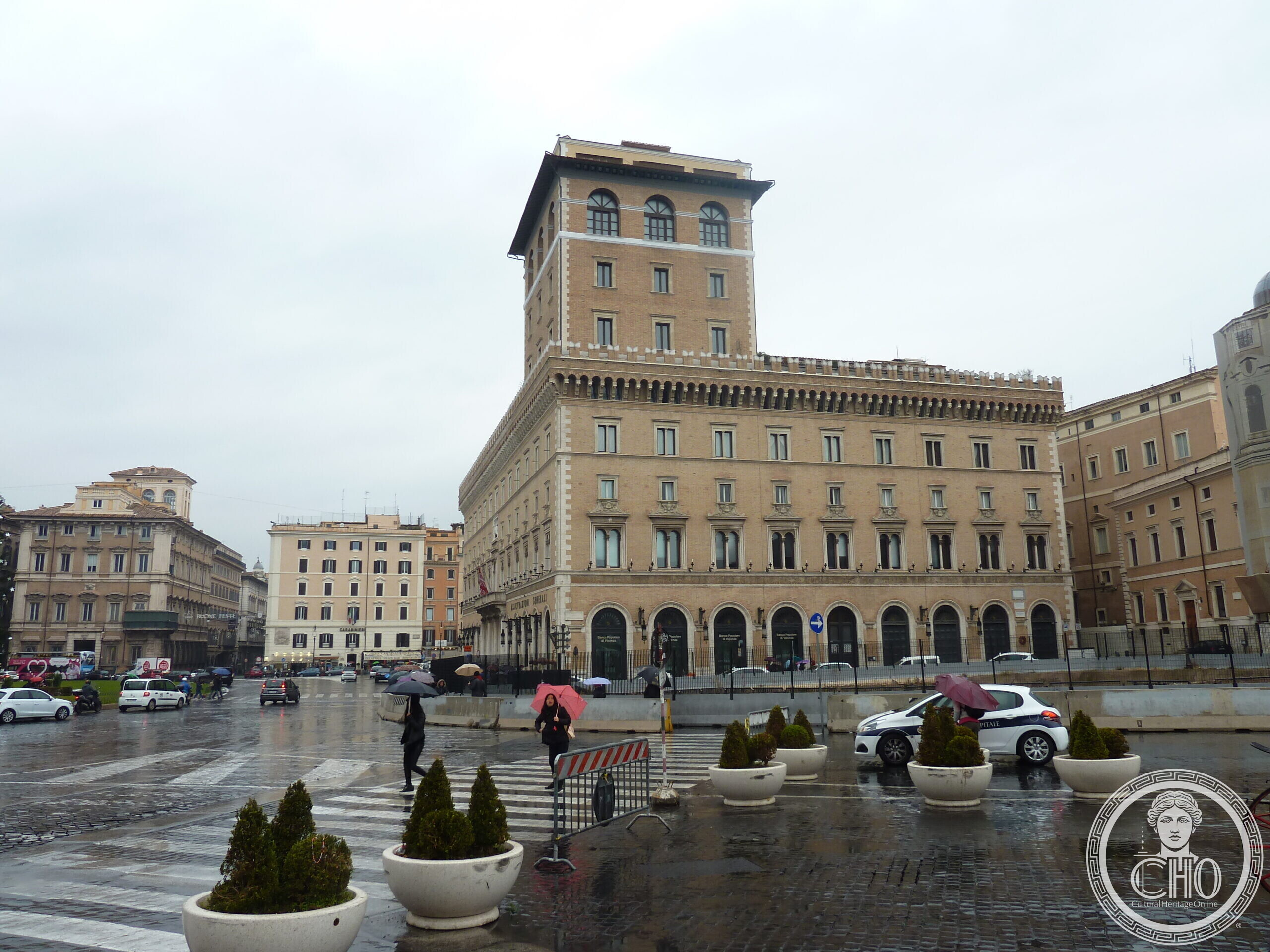 A long Renaissance palace front with three rows of windows and a crenellated top, a tall square tower at the corner, seen across a wet square with potted trees