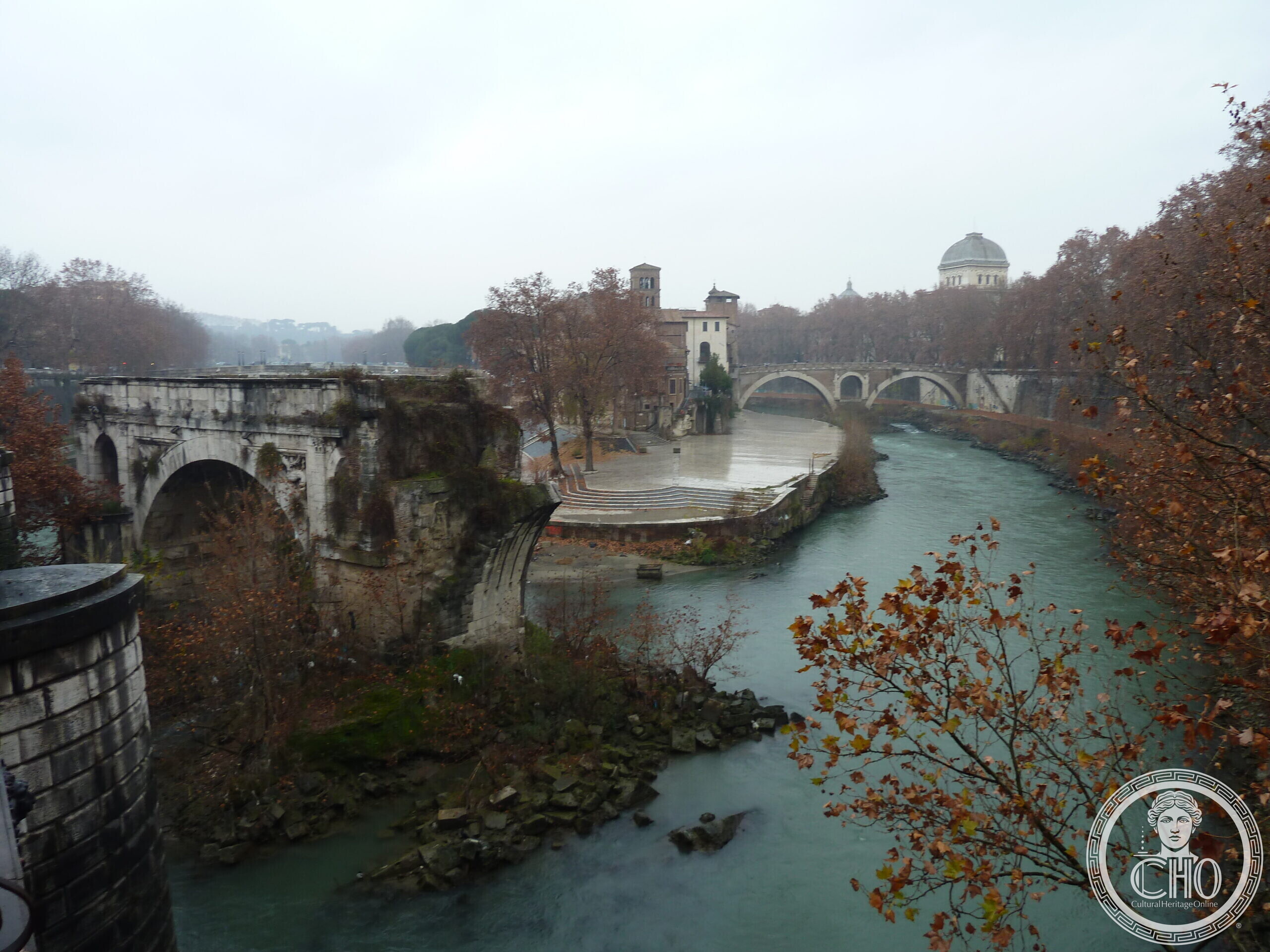 A single ancient stone arch standing alone in the green water of the Tiber, overgrown with plants, with the Tiber Island and a domed building in the misty distance