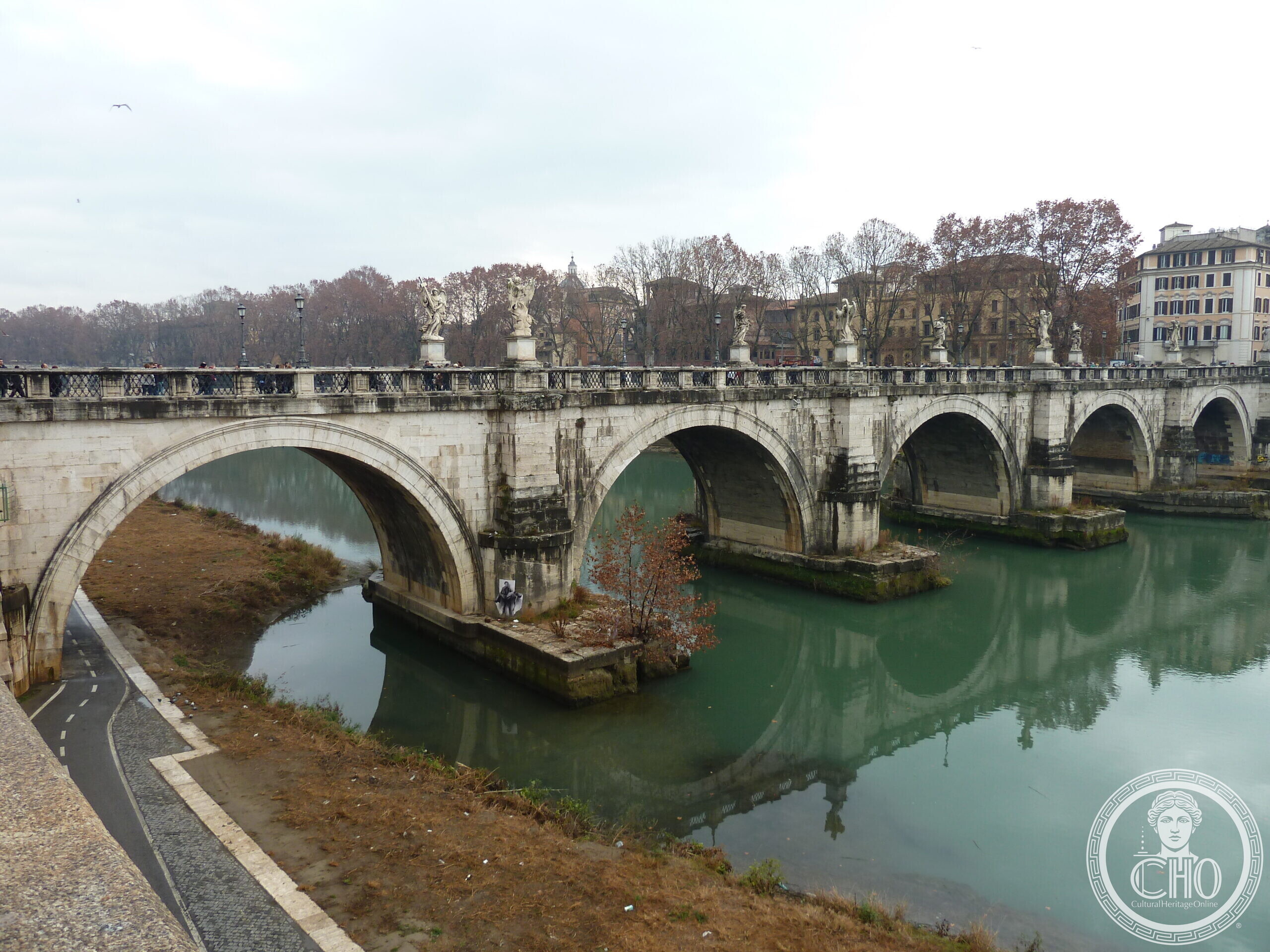 A stone bridge of several arches over a green river with statues standing on its parapets, seen from the embankment path on the near bank under a grey sky