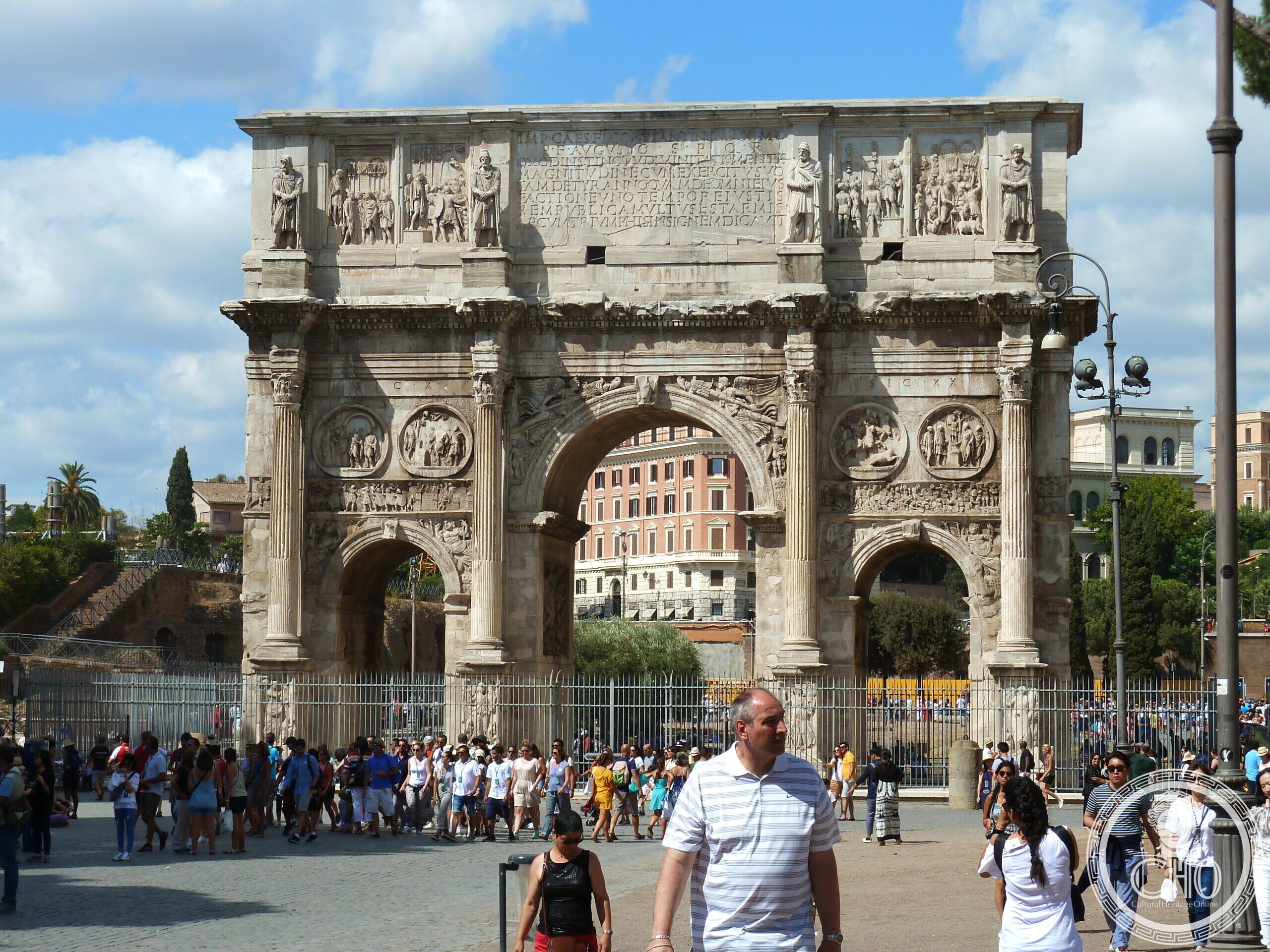 A large white marble triumphal arch with three passages, detached columns and statues on the attic, seen from the front across a crowd of visitors on a sunny day