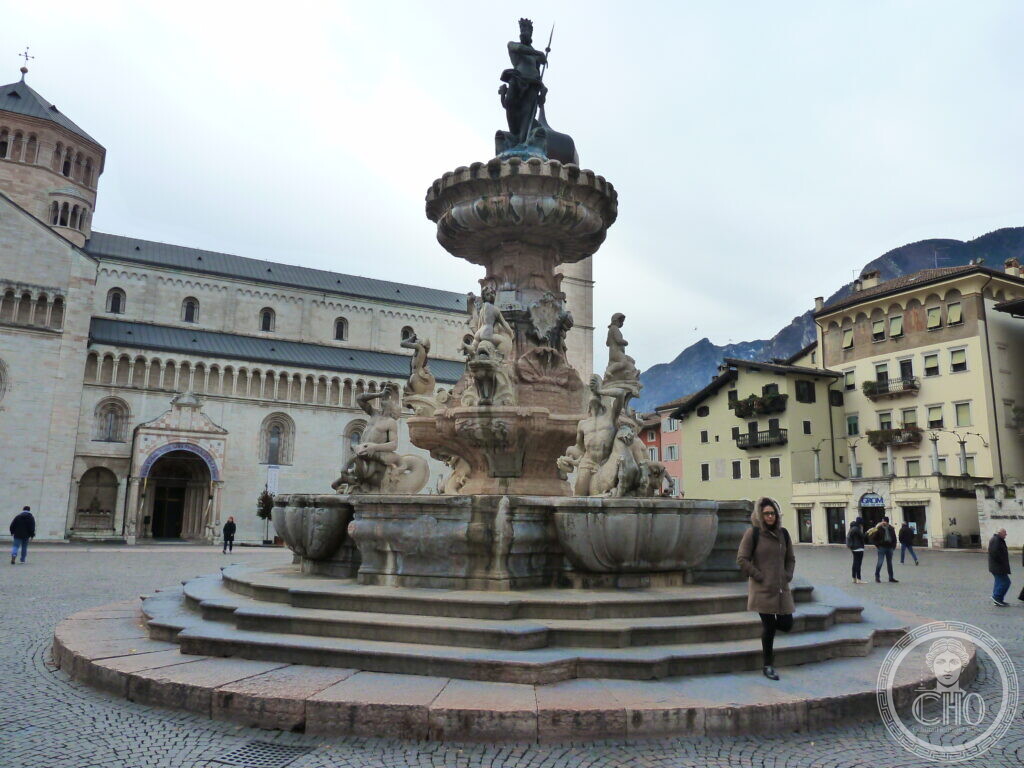 A large ornate stone fountain with a statue of Neptune on top stands in a paved square in front of a Romanesque church and colorful buildings.