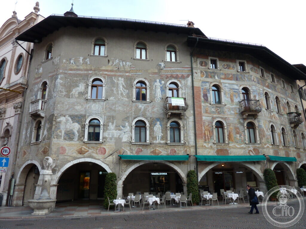 A large frescoed building with two arched arcades and green awnings on a paved street corner.