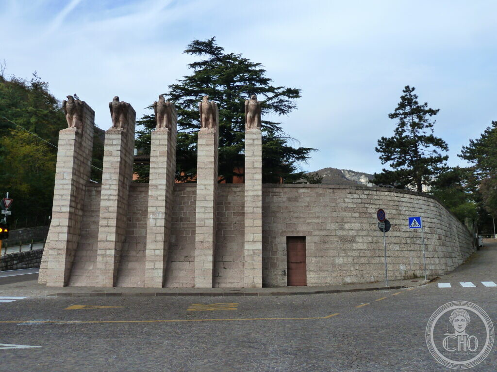 Five tall stone pillars topped with eagle sculptures stand in front of a curved stone wall beside a road.