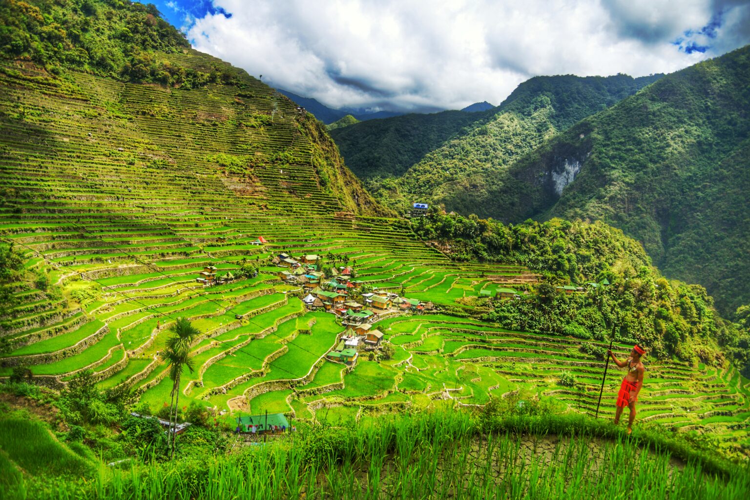 Wooden houses standing among flooded rice terraces at Banaue, with dense forest covering the ridge above the highest paddies